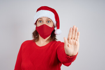 Fototapeta na wymiar An adult woman in a red protective mask, in a Santa hat on a gray background, shows a stop gesture. Christmas, New Year in quarantine