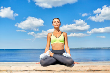 Fototapeta na wymiar fitness, sport and yoga concept - young woman meditating in lotus pose at seaside