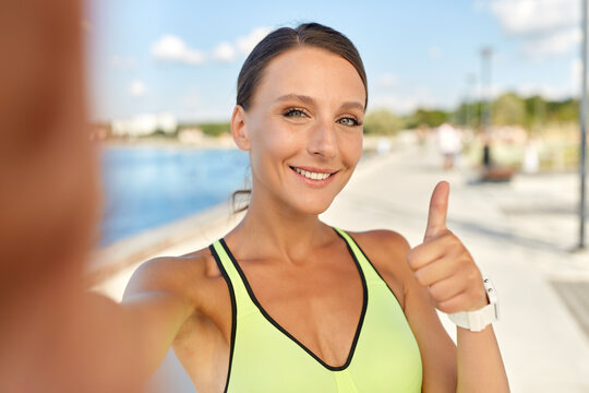people, sport and fitness concept - portrait of smiling young sporty woman taking selfie picture and showing thumbs up outdoors on sea promenade