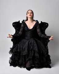 full length portrait of  woman wearing black gothic dress, sitting on a ornate black armchair....