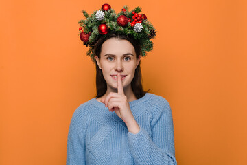 Positive female in handmade wreath, has toothy smile, makes hush gesture, wears winter sweater,...
