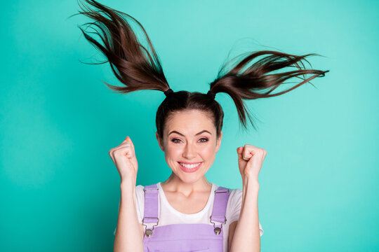 Photo of astonished lovely girl raise fists wind blow ponytails wear purple overall isolated teal color background