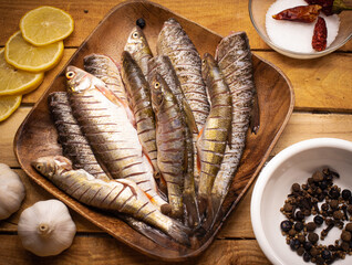 Flat lay fresh fish chub on a wooden squared brown plate on the wooden background decorated with lemon, spices, dried pepper, salt, and garlic