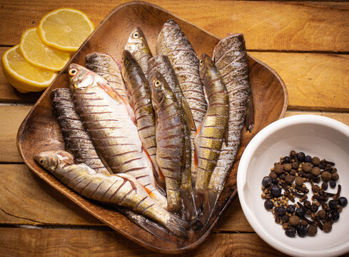 Flat lay fresh fish chub on a wooden squared brown plate on the wooden background decorated with lemon and spices in the white cup