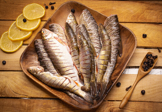 Flat lay fresh fish chub on a wooden squared brown plate on the wooden background decorated with lemon and spoon full of spices