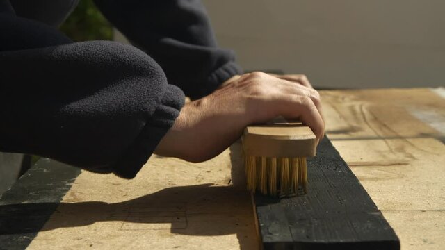 Pine wood plank with charred surface being scraped with wire brush by carpenter hands
