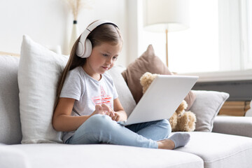 Online education of children. Sweet girl with headphones looking video lesson teacher conference laptop sitting on the couch at home.