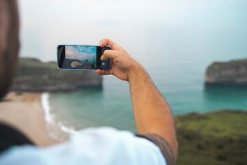 A man takes a picture with his cell phone, at the panorama. In the background, a beach, and the sea of spain.
