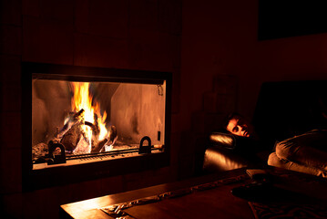 Teenager sleeping on the couch next to the fireplace with fire.