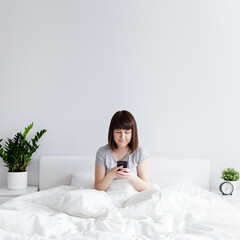 happy woman sitting on bed and using smart phone at home, copy space over white wall