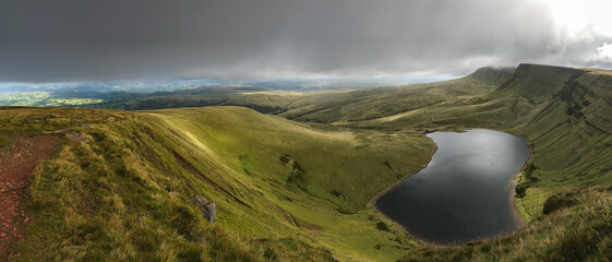 A panoramic view of the rolling green hills and mountains of the Brecon Beacons in Wales with a lake in the foreground and dark moody sky