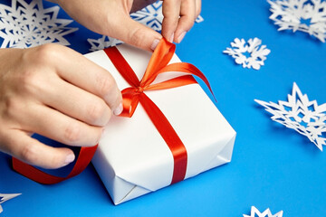 Hands tying red ribbon on white gift box on blue background