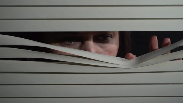 Close up front portrait from indoors of brunette woman pushing blinds at window with hands. Looking fearfully outside