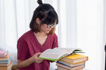 Serious Asian teen girl with eyeglasses pay attention reading book for examination or competition, university student read book at home get headache, woman casual with stack of books learning at home