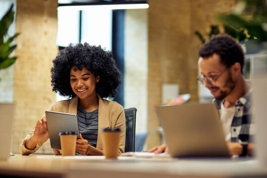 Two young happy multiracial business people sitting at the desk, using modern technologies while working together in coworking space