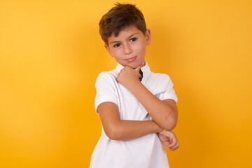 Thoughtful smiling Cute Caucasian little boy standing against yellow background keeps hand under chin, looks directly at camera, listens something with interest. Youth concept. © Jihan