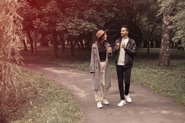 Fototapeta premium Young couple walking spring green city park, drinking hot coffee paper eco recycle cup. Two lovers husband wife spending time together, discuss news movie film black umbrella to shelter the rain.