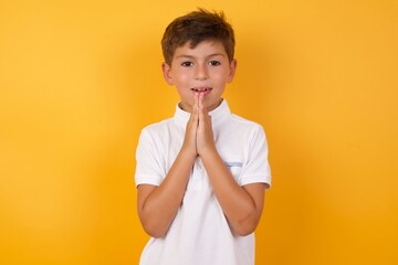Cute Caucasian little boy standing against yellow background  keeps palms together, has pleased...