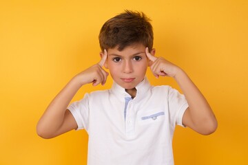 Cute Caucasian little boy standing against yellow background  concentrating hard on an idea with a serious look, thinking with both index fingers pointing to forehead.