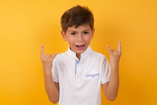 Born To Rock This World. Joyful Cute Caucasian Little Boy Standing Against Yellow Background  Screaming Out Loud And Showing With Raised Arms Horns Or Rock Gesture.