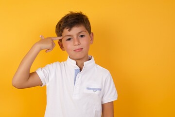 Unhappy Cute Caucasian little boy standing against yellow background  curves lips and makes suicide gesture,  shoots in temple with hand, tries kill himself.