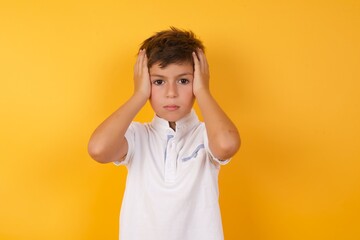 Frustrated Young handsome Caucasian little boy standing against yellow wall plugging ears with hands does not wanting to listen hard rock, noise or loud music.