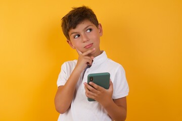 Thinking dreaming Young handsome Caucasian little boy standing against yellow background  using mobile phone and holding hand on face. Taking decisions and social media concept.