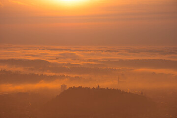 Amazing foggy sunrise over the city of Graz with Schlossberg hill and Church of the Sacred Heart of Jesus tower, in Styria region, Austria. Panoramic view from Plabutsch mountain on autumn morning.