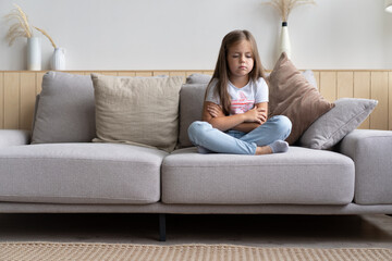 Beautiful unhappy sad little 6-year old girl sitting cross legged on couch in living room, feeling bored and lonely, being home alone while her parents spending all day at work
