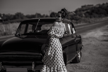 Gorgeous young woman wearing classy spot print off shoulders dress standing on the road leaning on her vintage car