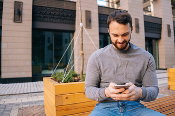Portrait of cheerful bearded young man wearing casual clothes using mobile phone while sitting on urban bench at empty European city street. Handsome male typing message on cellphone outdoors.