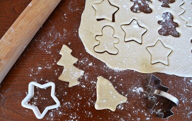 Christmas cookie dough stretched and cut with molds in the shape of a gingerbread man, stars, trees and bells with several cutters and a rolling pin next to it