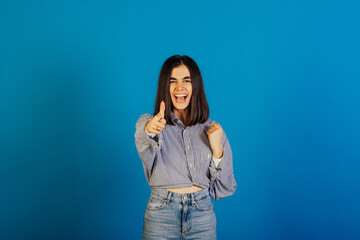 Young happy girl wearing stylish blue shirt and jeans look at camera show cool gesture. Cheerful girl showing thumbs up over blue background. 