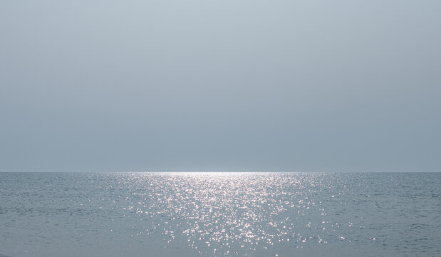 A calm sea with light ripples and sun glare in the center and a cloudness serene sky. A clear line of the horizon in the seascape, gray-blue tones.