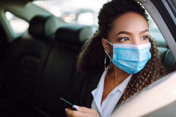 Fototapeta na wymiar Woman wearing a medical sterile mask in taxi car on a back seat looking out of window checking her cell phone. Safety and pandemic concept. Covid-2019.