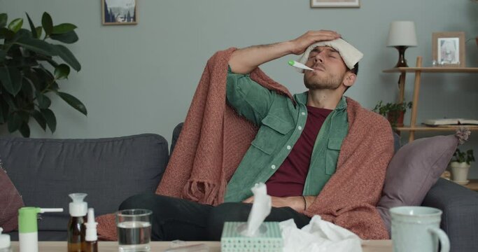 Ill young man measuring body temperature while sitting on sofa at home. Bearded guy in 30s with termometr in mouth holding cold compress on his forehead. Concept of illness and medicine