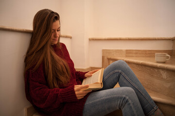 pretty young woman reading a book and drinking coffee happily, sitting on some stairs