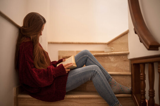 pretty young woman reading a book and drinking coffee happily, sitting on some stairs
