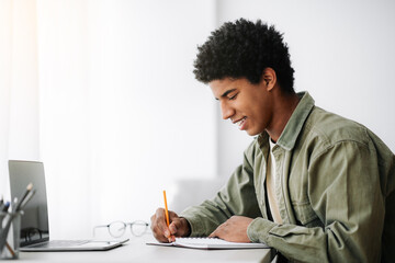 Web-based training concept. Black male student writing in notebook in front of laptop at home,...