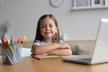 Portrait of happy small pupil learning at home. Smiling little child girl enjoying doing lessons in living room. Smart kid schoolgirl looking at camera, studying remotely online.