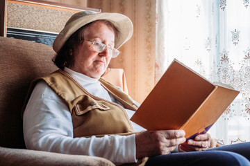 Fototapeta na wymiar Elderly woman with a hat on, reading a book by the window. Afternoon light