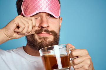 Man with pink sleep mask on his face and a cup of tea in his hands cropped view close-up blue background