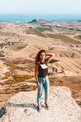 A young beautiful woman with long hair and jeans stands on a rock in the Koktebel valley in Crimea. The landscape of sandy hilly desert.