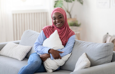 Home Comfort. Portrait Of Smiling Muslim Woman In Hijab Sitting On Sofa And Cuddling Pillow, Relaxing In Living Room