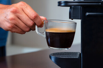 Close up of hand picking up a cup of espresso of capsule coffee machine at home. Concept of coffee break.