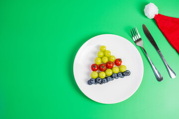 Christmas tree of organic vegetable and fruit; Blueberries, grape, Strawberry and tomato with fork and knife on green background. Merry Xmas and happy New Year party, New You, Healthy food concept