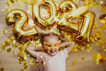 Cheerful child girl in a mask and with the numbers 2021 rejoices at the confetti flying from above, top view. New Year celebration concept. Holidays at home