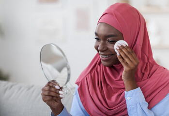 Attractive black muslim woman cleaning her face with cotton pad at home