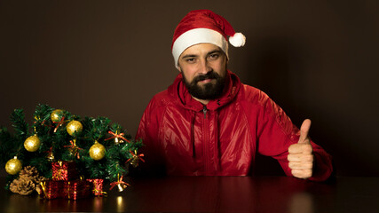 Young modern santa claus is sitting at a table near the tree. Emotional portrait. Thumds up