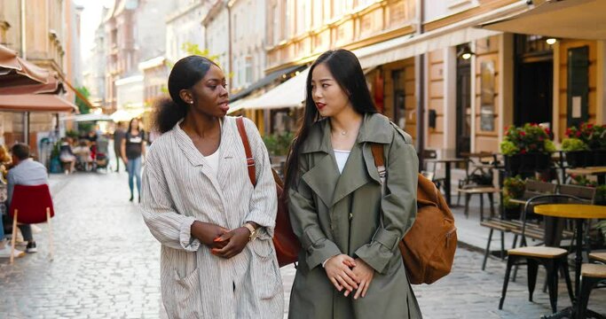 Mixed-races females best friends walking the street, talking and sharing secrets. Multi ethnic young women chatting and strolling outside. Cheerful Asian and African American girls gossiping. Gossips.